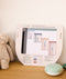 A sage green portable white noise machine with a white base rests on a wooden surface next to a printed What to Wear guide showing room temperatures and appropriate sleepwear.