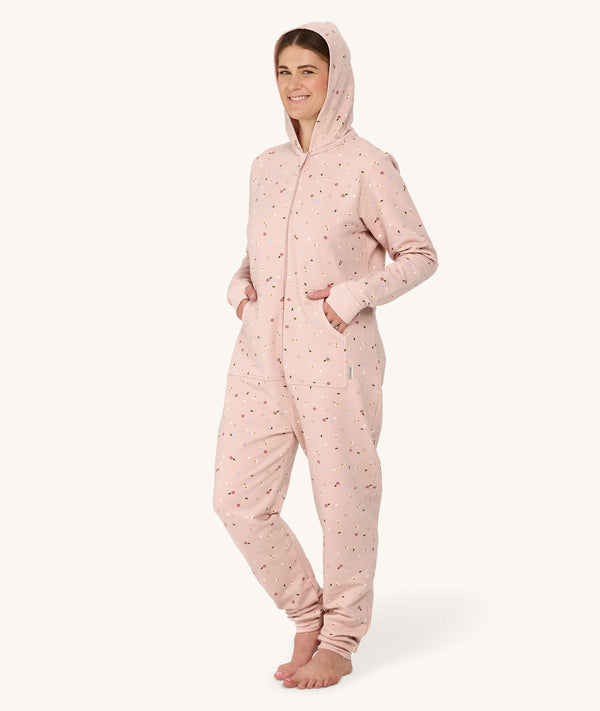 Barefoot woman standing against a white background wearing a hooded light pink floral Adult Sleep Onesie with her hands in the pockets.