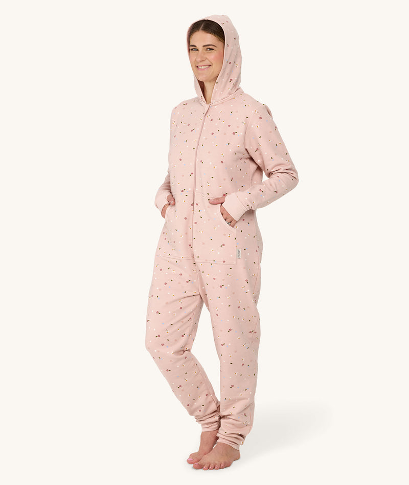 Barefoot woman standing against a white background wearing a hooded light pink floral Adult Sleep Onesie with her hands in the pockets.