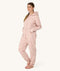 Woman standing barefoot against a white background wearing a light pink hooded Adult Sleep Onesie with floral pattern, with her hands resting in front pockets.