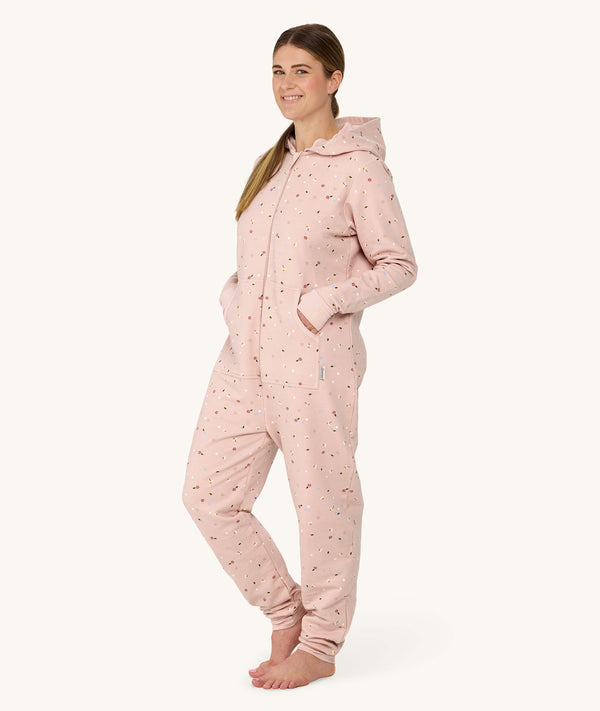 Woman standing barefoot against a white background wearing a light pink hooded Adult Sleep Onesie with floral pattern, with her hands resting in front pockets.