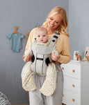 Mum wearing a baby in carrier. Baby wears an ergoPouch Sleep Suit Bag in Fawn.