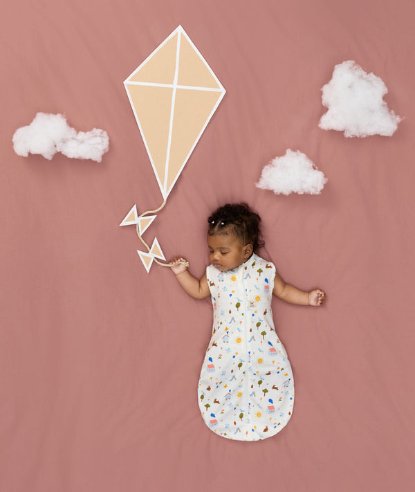 Newborn asleep in ergoPouch Cocoon Swaddle Bag 0.2 TOG Daydream, arms out sleeping, with a geometric kite and clouds