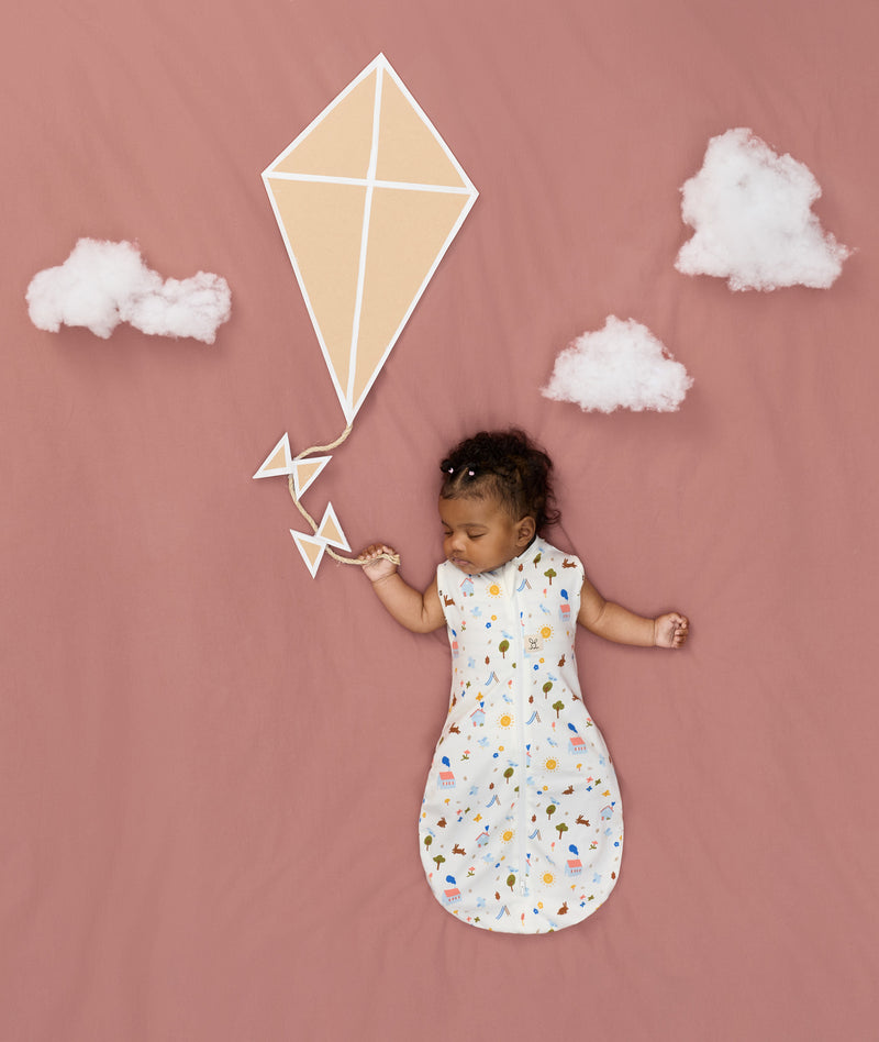Newborn asleep in ergoPouch Cocoon Swaddle Bag 0.2 TOG Daydream, arms out sleeping, with a geometric kite and clouds