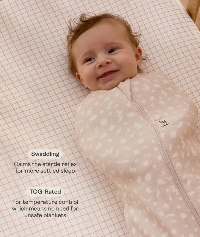 Smiling baby wrapped in a Cocoon Swaddle Bag lying on a grid-patterned sheet with text highlighting  swaddling benefit and TOG rating feature.
