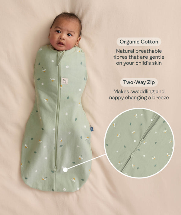 Baby swaddled in a green floral Cocoon Swaddle Bag, lying on a cream-coloured blanket with text callouts highlighting organic cotton and two-way zip features.