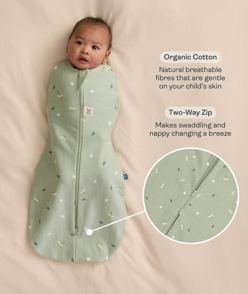 Baby swaddled in a green floral Cocoon Swaddle Bag, lying on a cream-coloured blanket with text callouts highlighting organic cotton and two-way zip features.