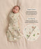 A baby lying on a light background wearing an animal-patterned Cocoon Swaddle Bag with product feature callouts: organic cotton and two-way zip. A circular inset zooms in on the double-zipper detail.