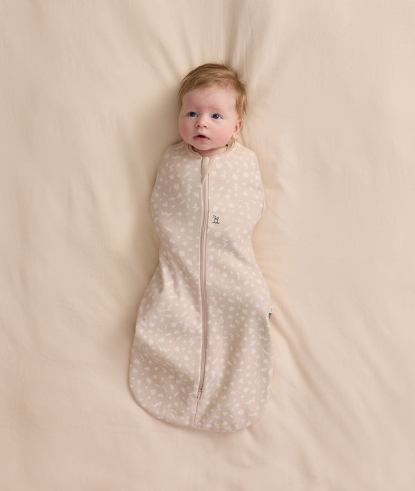 An overhead view of a baby swaddled in a patterned beige Cocoon Swaddle Bag, lying flat on a cream-colored blanket.