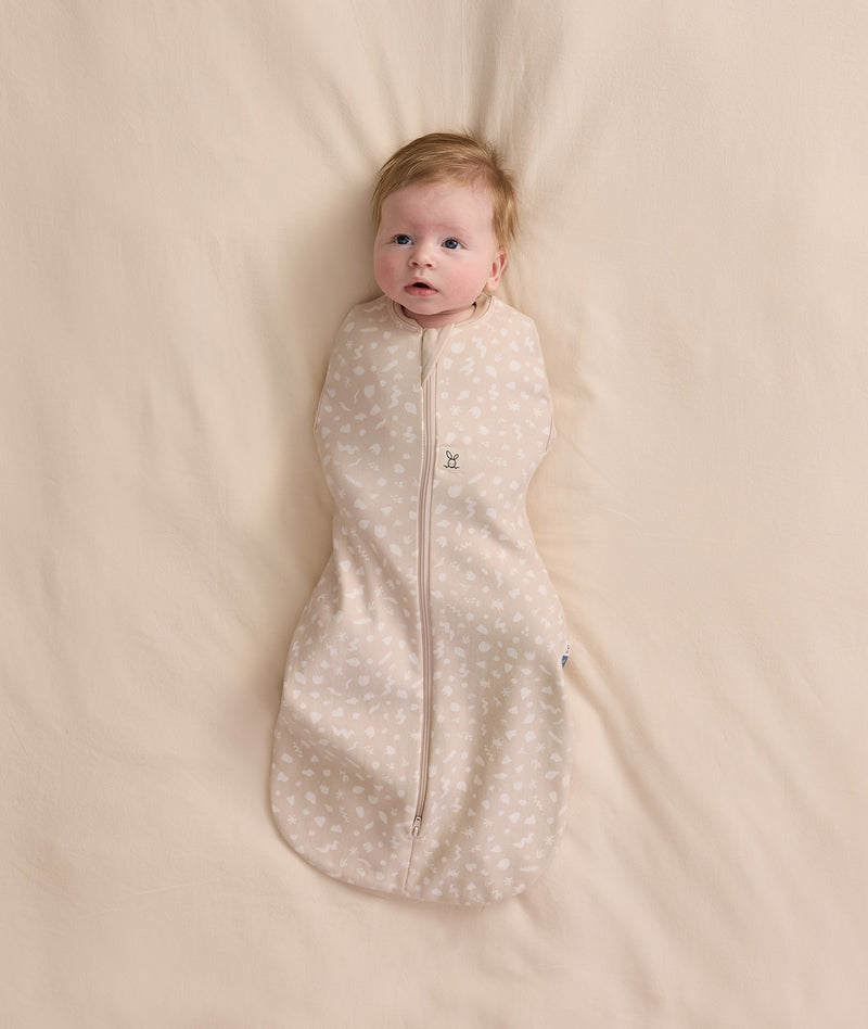 An overhead view of a baby swaddled in a patterned beige Cocoon Swaddle Bag, lying flat on a cream-colored blanket.