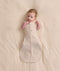Baby lying on a cream blanket, wearing a patterned Cocoon Swaddle Bag 1.0 TOG with both arms out