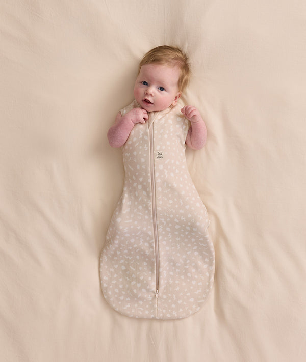 Baby lying on a cream blanket, wearing a patterned Cocoon Swaddle Bag 1.0 TOG with both arms out