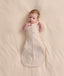 Baby lying on a cream blanket, wearing a patterned Cocoon Swaddle Bag 1.0 TOG with both arms out