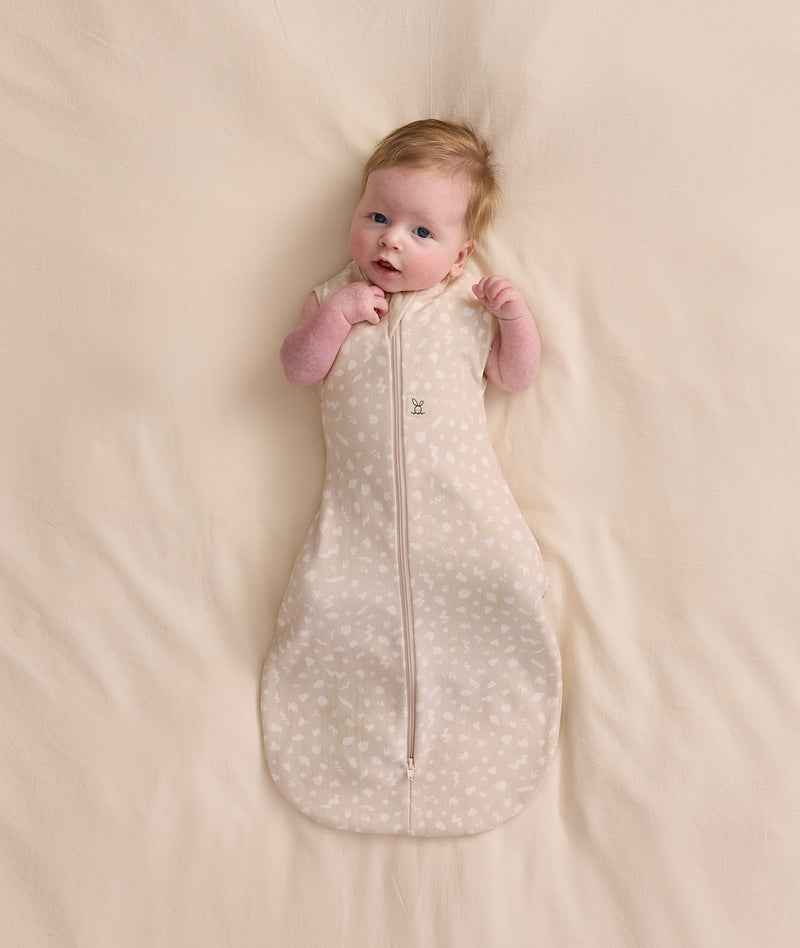 Baby lying on a cream blanket, wearing a patterned Cocoon Swaddle Bag 1.0 TOG with both arms out