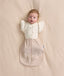 An overhead view of a baby lying flat on a cream-colored sheet, wearing a patterned beige Cocoon Swaddle Bag with a Butterfly Cardi layered over top.