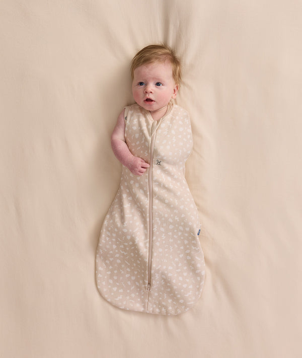 An overhead view of a baby lying flat on a cream-colored sheet, wearing a patterned Cocoon Swaddle Bag with one arm extended out.