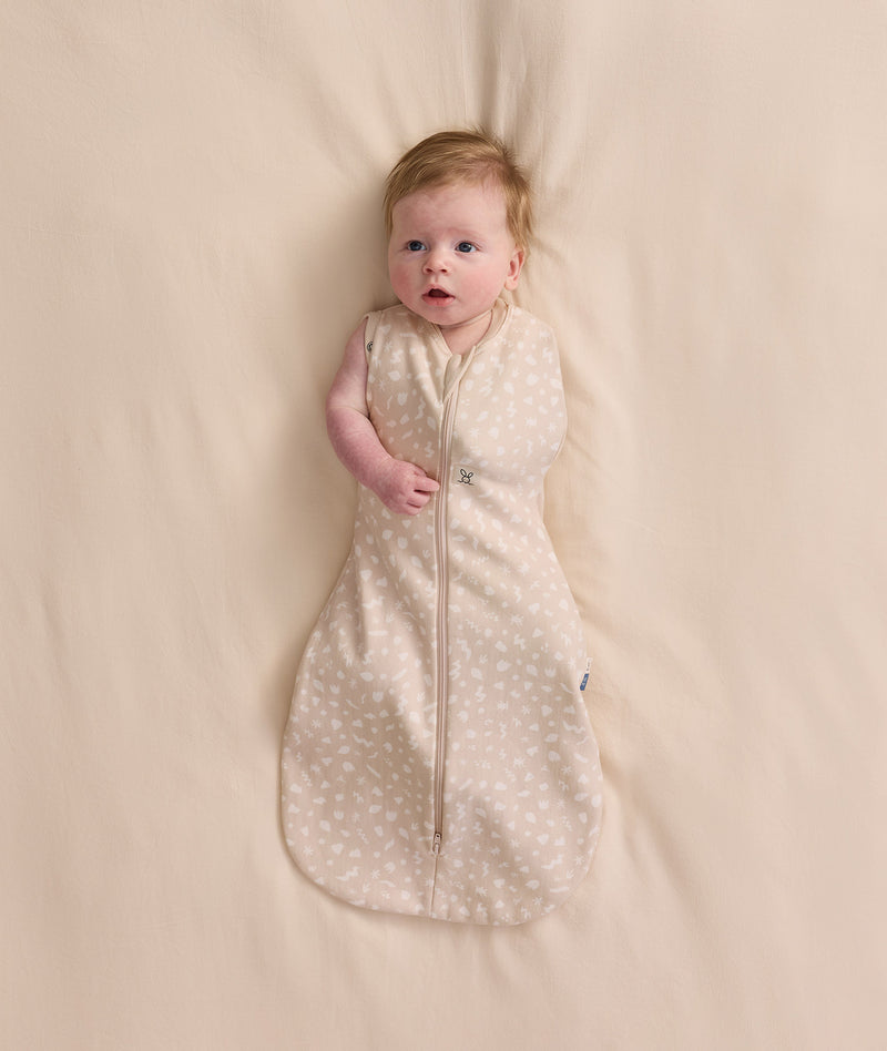 An overhead view of a baby lying flat on a cream-colored sheet, wearing a patterned Cocoon Swaddle Bag with one arm extended out.