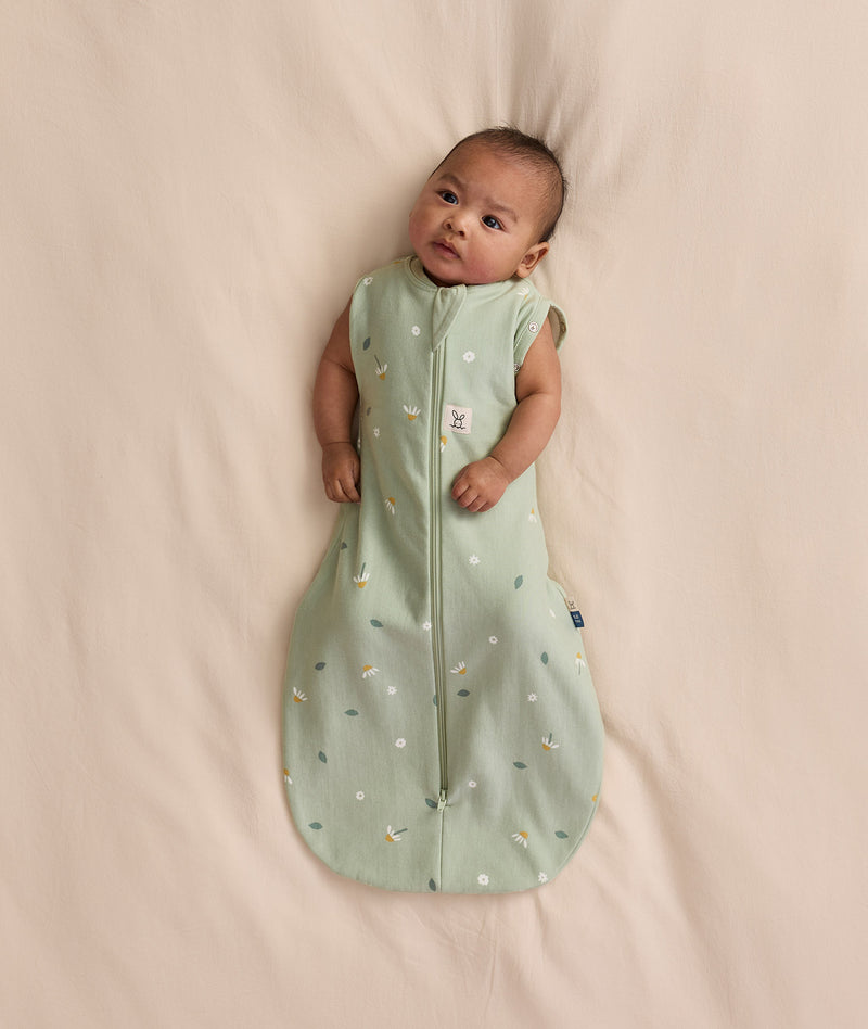 Baby lying on a cream blanket, wearing an ergoPouch Cocoon Swaddle Bag 1.0 TOG with both arms out.