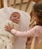 A young girl in pink pajamas reaching into a wooden crib to gently place her hand on a swaddled newborn. The baby is swaddled in a safari-print Cocoon Swaddle Bag.