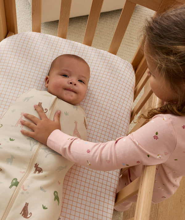 A young girl in pink pajamas reaching into a wooden crib to gently place her hand on a swaddled newborn. The baby is swaddled in a safari-print Cocoon Swaddle Bag.