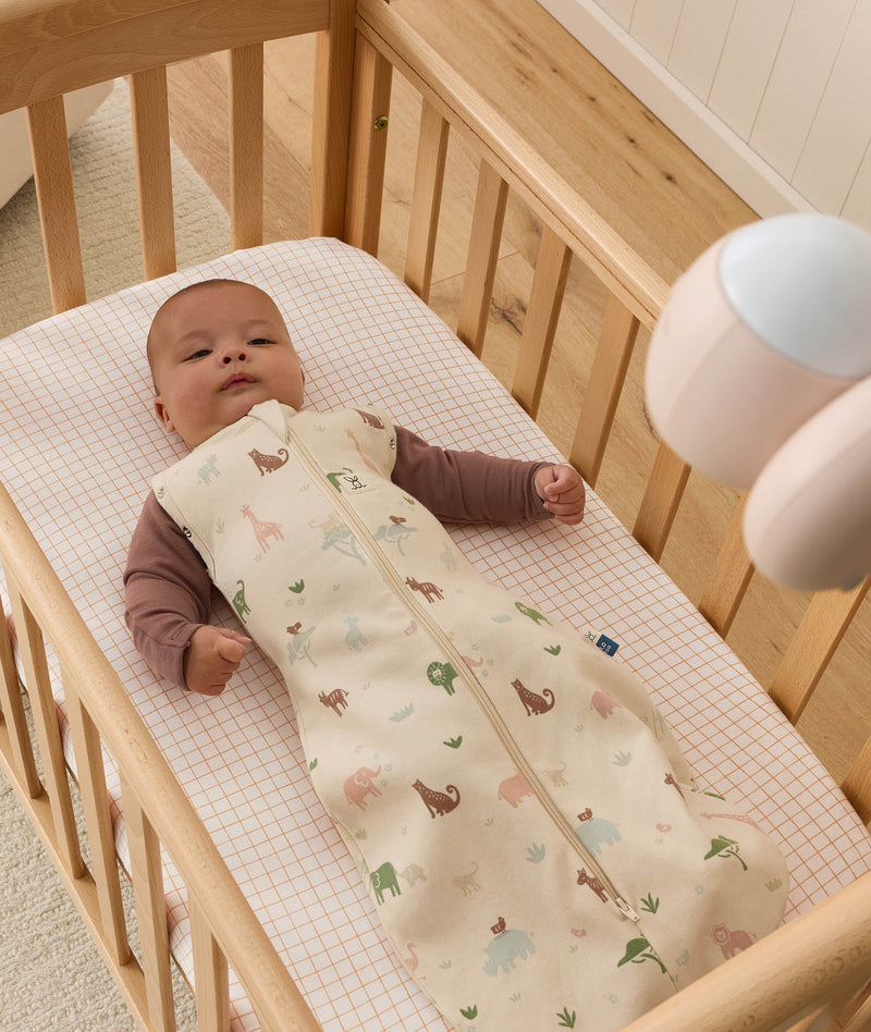 A baby wearing a brown long-sleeve romper and a safari-print Cocoon Swaddle Bag lying in a wooden crib.