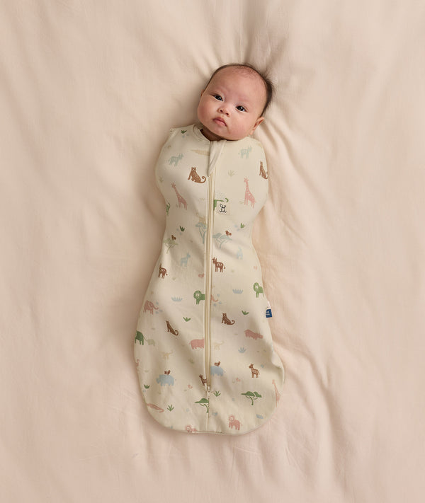Baby lying on a cream blanket swaddled in a safari-patterned Cocoon Swaddle Bag 1.0 TOG.
