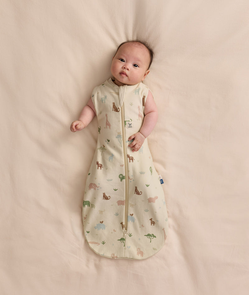 Baby lying on a cream-colored blanket wearing a Cocoon Swaddle Bag 1.0 TOG with both arms out.