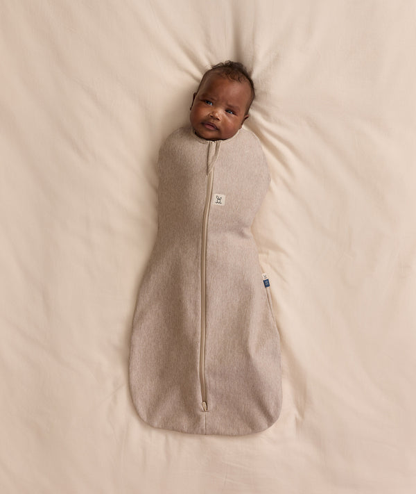 Baby lying on a cream blanket, swaddled in an ergoPouch Cocoon Swaddle Bag 1.0 TOG Taupe Marle