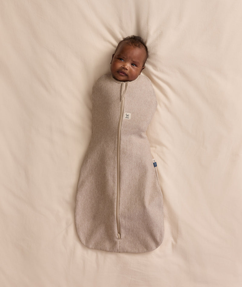 Baby lying on a cream blanket, swaddled in an ergoPouch Cocoon Swaddle Bag 1.0 TOG Taupe Marle