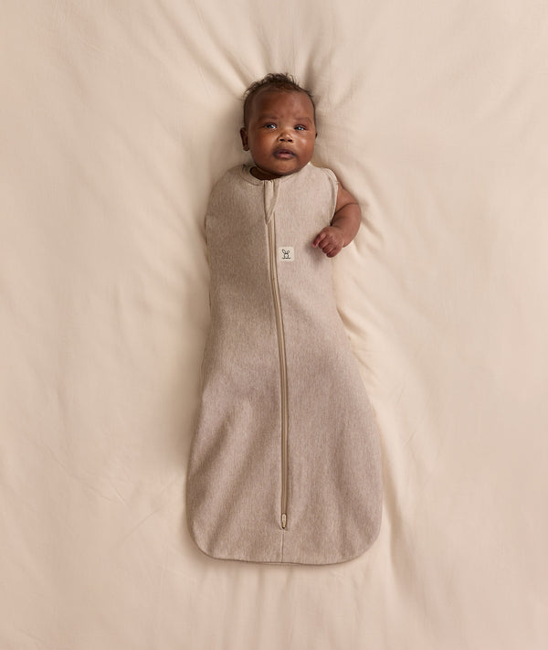 Baby lying on a cream-coloured blanket, wearing an ergoPouch Cocoon Swaddle Bag with one arm extended out.