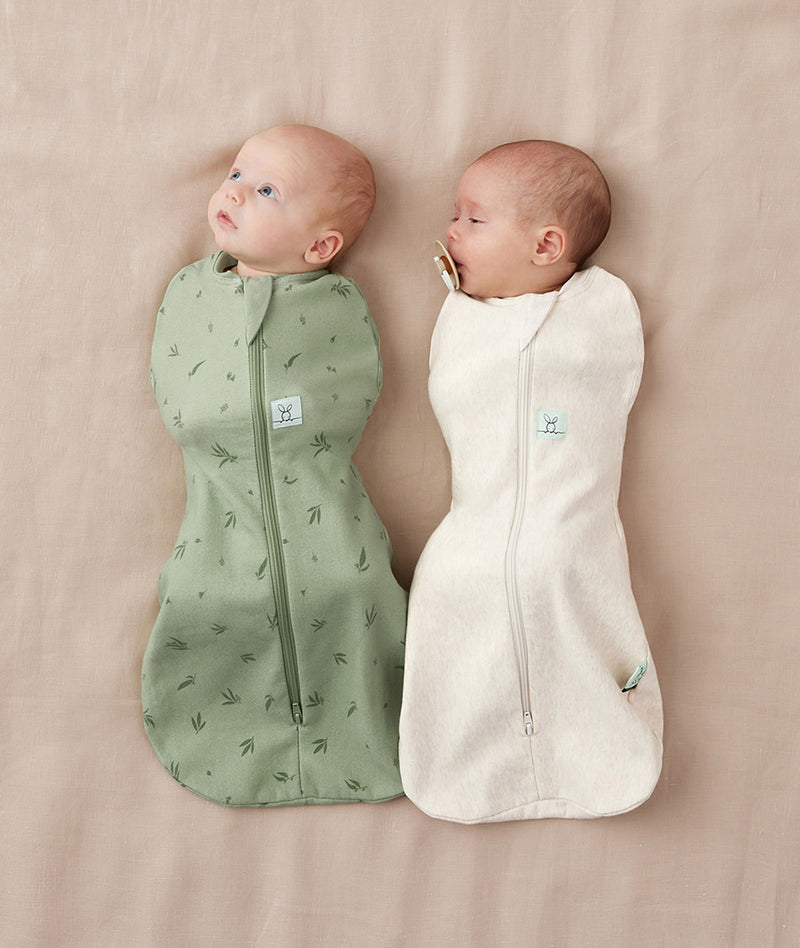 Two newborn babies lying side by side, being swaddled in an ergoPouch Cocoon Swaddle Bag 1.0 TOG in Willow and Oatmeal Marle.