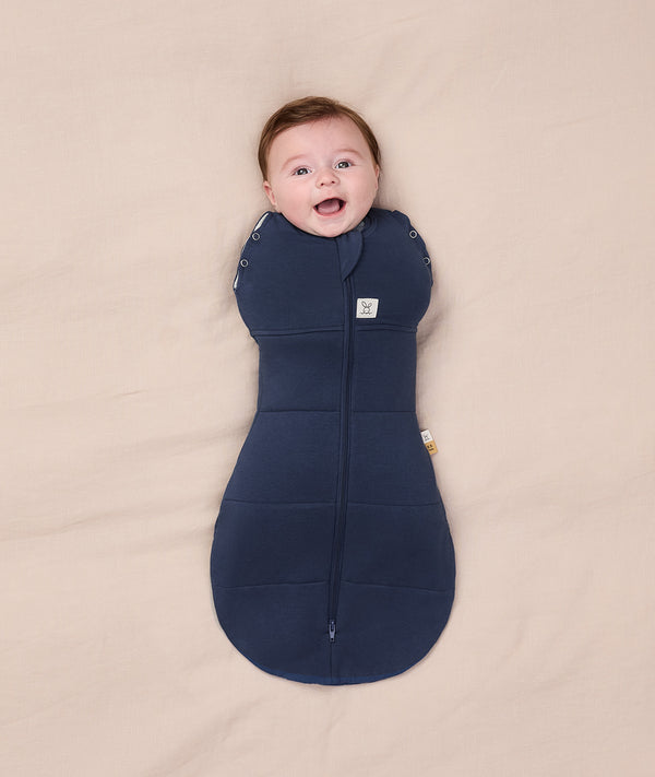 A smiling baby wrapped  in a dark blue Cocoon Swaddle Bag 2.5 TOG on a cream-coloured blanket