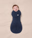 A smiling baby wrapped  in a dark blue Cocoon Swaddle Bag 2.5 TOG on a cream-coloured blanket