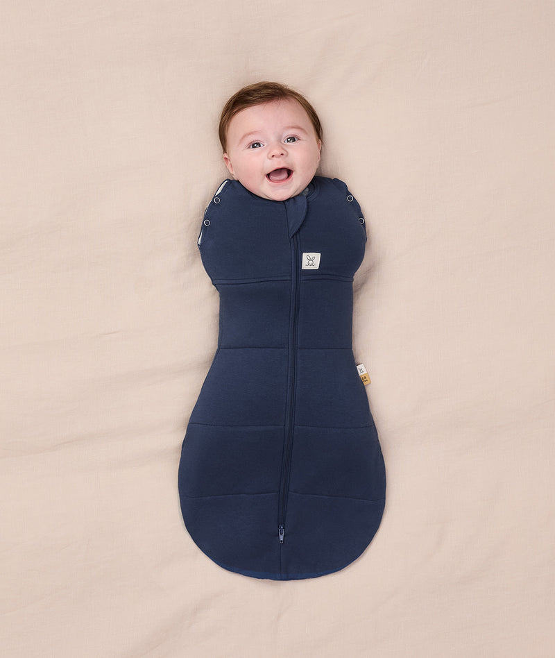 A smiling baby wrapped  in a dark blue Cocoon Swaddle Bag 2.5 TOG on a cream-coloured blanket