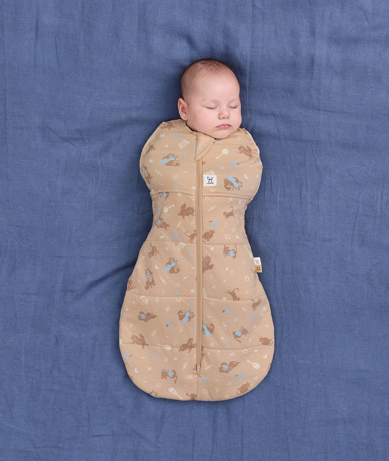A newborn sleeping peacefully while wrapped snugly in a tan, patterned Cocoon Swaddle Bag 2.5 TOG on a blue fabric background.