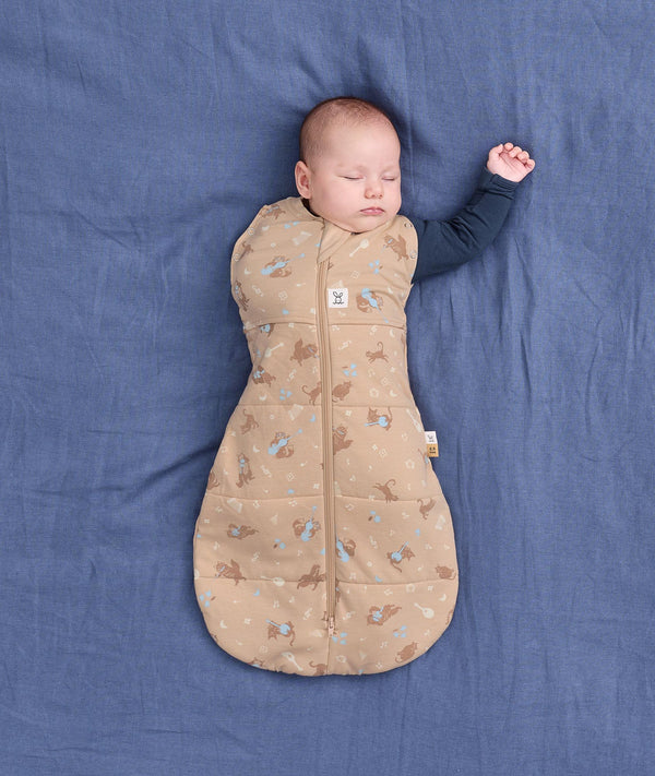 Sleeping baby wearing a tan patterned Cocoon Swaddle Bag with their one arm stretched out.