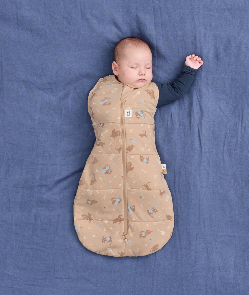 Sleeping baby wearing a tan patterned Cocoon Swaddle Bag with their one arm stretched out.