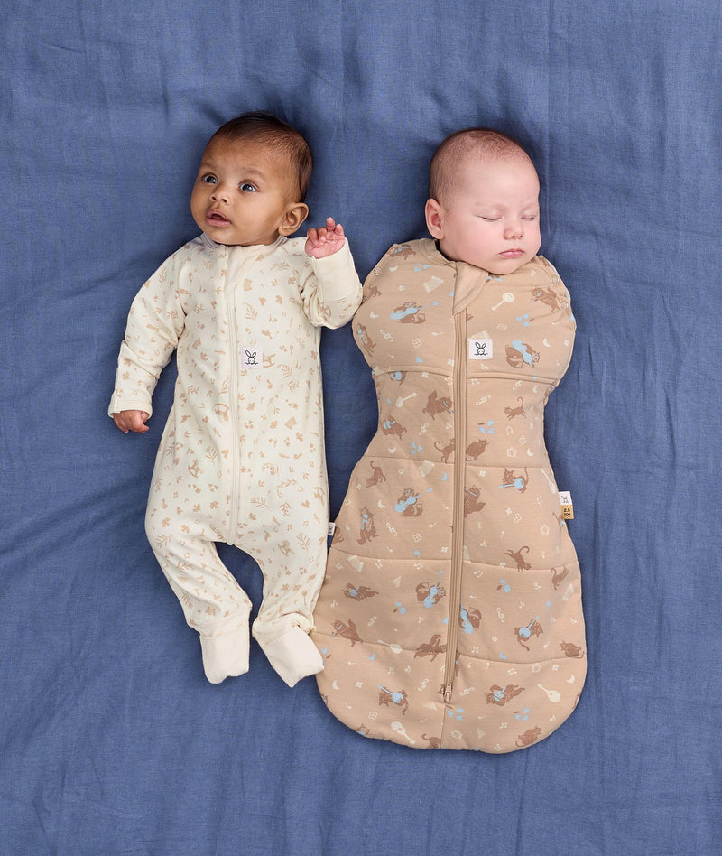 Two babies lying on a blue fabric background wearing ergoPouch sleepwear. One wearing a Long Sleeve Layer in Lullaby and the other one is wrapped in a 2.5 TOG Cocoon Swaddle Bag Melody.
