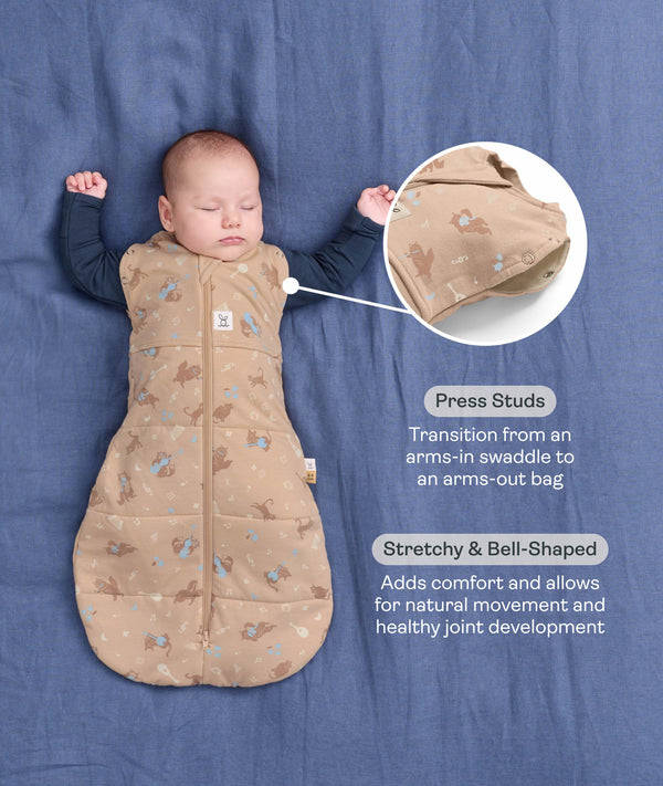 Product infographic showing a baby sleeping arms-out in a Cocoon Swaddle Bag on a blue background, highlighting armhole press studs and a bell-shaped design.