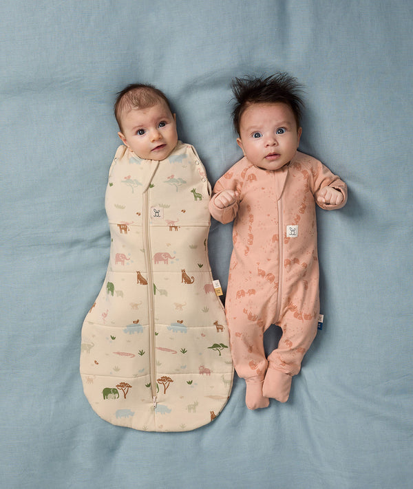 Two babies are lying side by side wearing ergoPouch Cocoon Swaddle Bag Savannah and Long Sleeve Layer Elephant Parade.