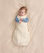 Baby lying on a cream-colored fabric background wearing a Cocoon Swaddle Bag with a blue Long Sleeve Layer underneath.
