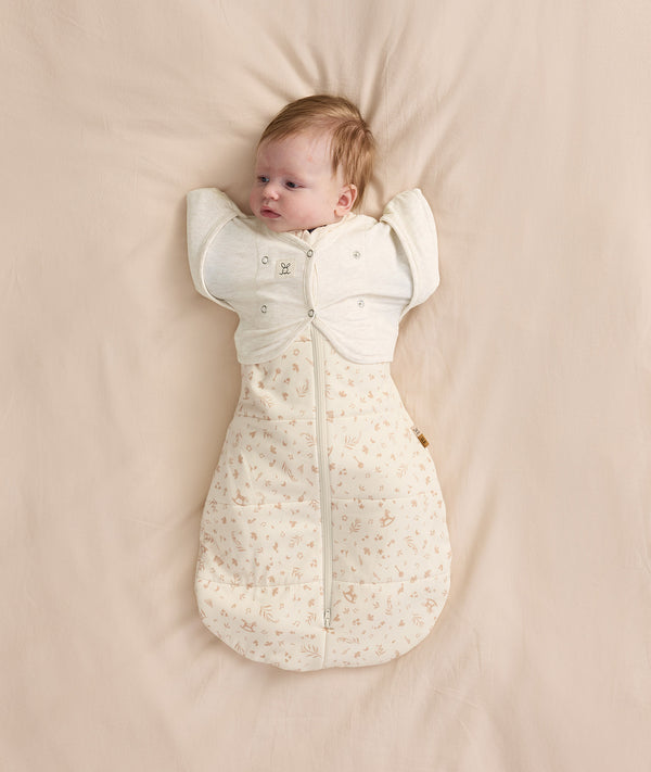 Newborn baby wearing a cream patterned Cocoon Swaddle Bag with Butterfly Cardi layered over top for hands up sleeping.