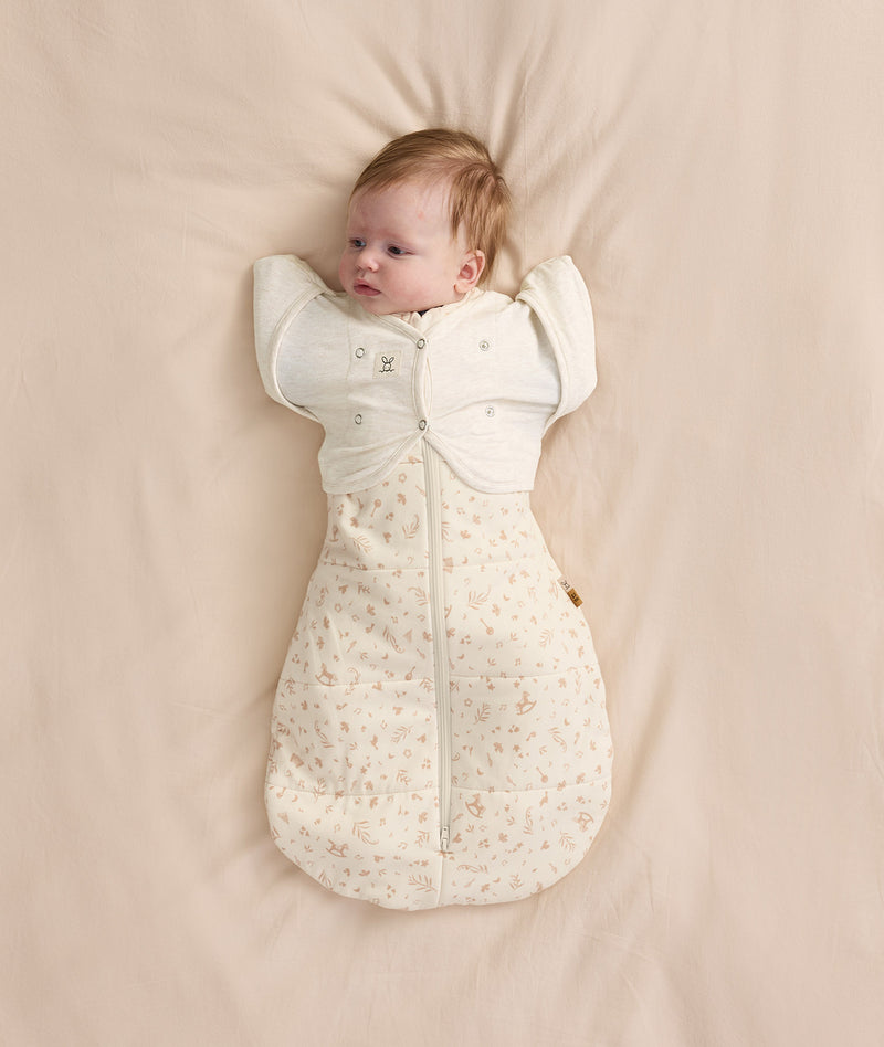 Newborn baby wearing a cream patterned Cocoon Swaddle Bag with Butterfly Cardi layered over top for hands up sleeping.