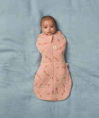 Baby lies on a soft blue blanket, wearing an ergoPouch Cocoon Swaddle Bag with its arms inside.