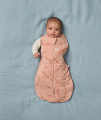 Baby lies on a soft blue blanket, wearing an ergoPouch Cocoon Swaddle Bag with its one arm outside.