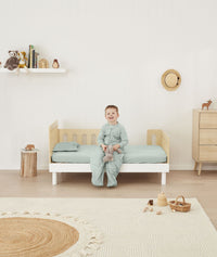 Toddler sitting on the side of a bed made with ergoPouch Organic Fitted Sheet Sage