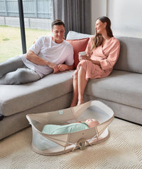 Parents relaxing on sofa, smiling at baby laying in ergoPouch Easy Sleep Portable Crib Ecru