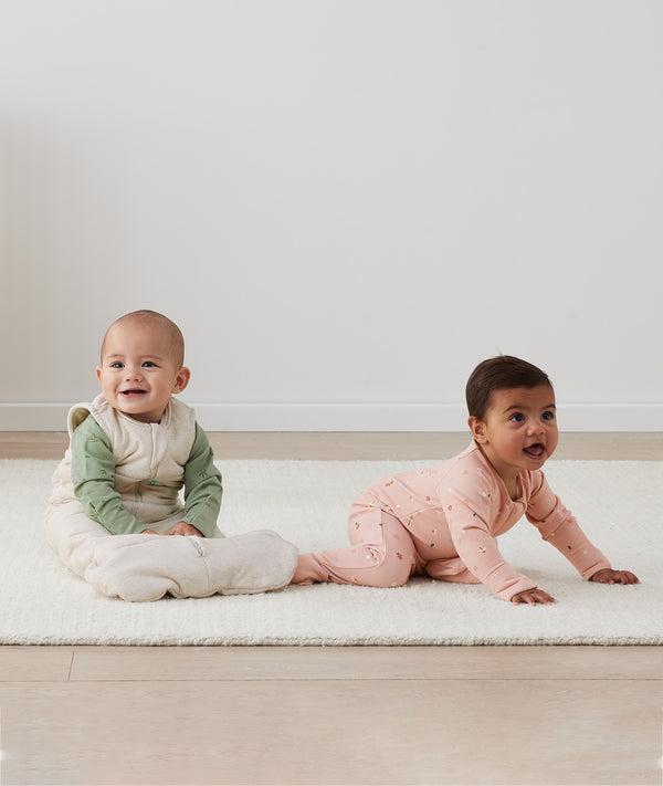 Two infants on the floor, one wearing an ergoPouch Long Sleeve Layer 1.0 TOG Daisies, and the other layered in a warmer Long Sleeve Layer 1.0 TOG Willow with a Sleeping Bag 2.5 TOG Oatmeal Marle overtop for warmth. 