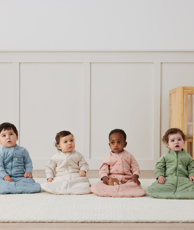 Four babies sitting on a white floor wearing ergoPouch Sleeping Bag Sleeved in 2.5 and 3.5 TOGs in heritage colours: Dragonflies, Oatmeal Marle, Daisies and Willow.