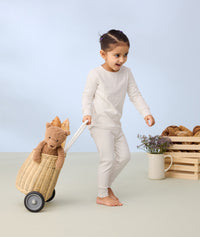 A young child wearing a long-sleeve pyjamas is happily pulling a small wicker trolley filled with a plush teddy bear and toy baguettes.